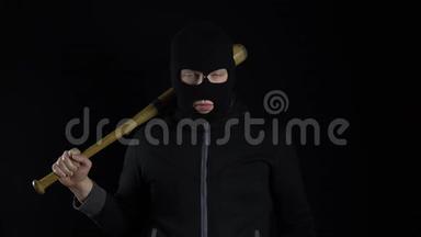 一个戴着巴拉克拉瓦面具的人正拿着棒球棒站着。 一个强盗拿着棒球棒站在黑色的背景上